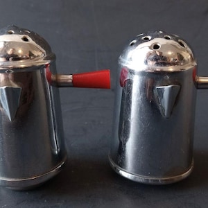 May include: Two vintage chrome salt and pepper shakers with red handles. Each shaker has a conical shape with a silver metal body and a series of holes on the top. A small, silver triangle is on the front of each shaker.