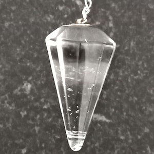 May include: A clear, faceted crystal pendulum suspended by a silver chain. The pendulum has a pointed, conical shape and is designed for divination or spiritual practices. The crystal is transparent, allowing light to pass through, and is set against a dark, textured background.