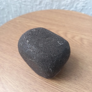 May include: A dark gray, porous stone with a rounded shape. The stone is resting on a light brown wooden surface. The stone's texture appears rough and uneven, with small holes and imperfections.