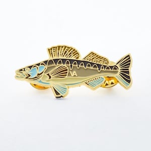 Op de afbeelding: Gouden metalen pin van een gestileerde vis met zwarte en bruine markeringen en blauwe accenten. De vis heeft een rugvin en een staartvin. De pin heeft de letters "VA" aan de zijkant.