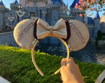 May include: A pair of beige mouse ears with a brown bow and gold glitter. The ears are embroidered with the words "Welcome" and "Happily Ever After".
