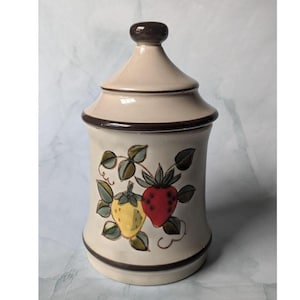 May include: A beige ceramic jar with a lid, featuring a brown trim and a decorative strawberry design. The jar has a cylindrical shape with a slightly flared base and a domed lid. The strawberries are red and yellow with green leaves.