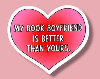 May include: A heart-shaped sticker with a white border and a gradient pink and red center. The text "MY BOOK BOYFRIEND IS BETTER THAN YOURS" is printed in black, bold letters.