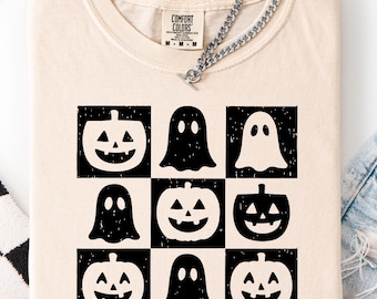 May include: Cream-colored t-shirt featuring a grid of black and white Halloween-themed graphics, including jack-o'-lanterns and ghosts. The shirt has a crew neck and short sleeves. A silver chain necklace is draped across the shirt. The shirt is made of a soft, comfortable material.