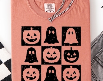May include: A coral-colored t-shirt featuring a black graphic design of pumpkins and ghosts in a grid pattern. The shirt has a crew neck and a silver chain necklace. The Comfort Colors brand tag is visible. Halloween-themed apparel.