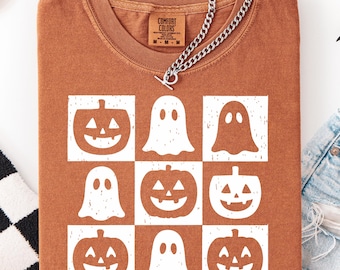 May include: A burnt orange t-shirt featuring a grid of white Halloween-themed graphics. The design includes jack-o'-lanterns and ghosts. A silver chain necklace is draped across the shirt. The shirt is made of a soft material.