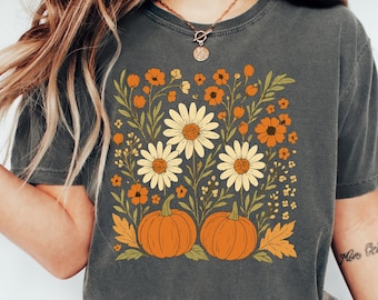 May include: Dark gray t-shirt featuring a floral design with large white daisies, orange flowers, and two orange pumpkins. The design includes green leaves and is a fall-themed graphic.