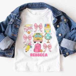 May include: White t-shirt with a colorful school-themed graphic, including books, bows, a school bus, a globe, and a backpack. The name "REBBECA" is printed in pink. The shirt is paired with a blue denim jacket.