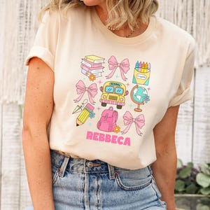 May include: Beige t-shirt with a colorful school-themed graphic. The design includes a school bus, books, pencils, a backpack, bows, and a globe. The name "REBBECA" is printed in pink letters.