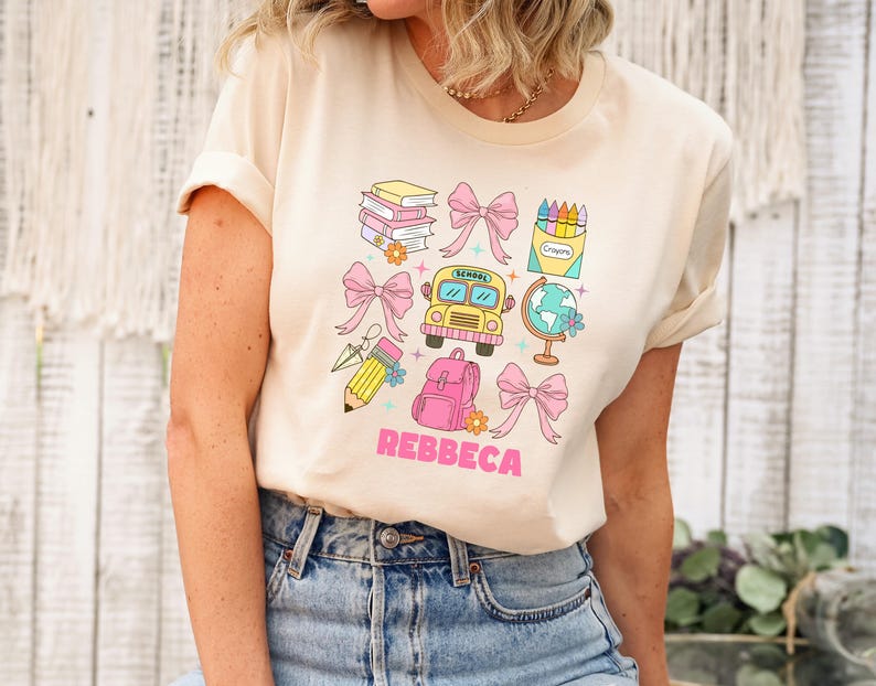 May include: Beige t-shirt with a colorful school-themed graphic. The design includes a school bus, books, pencils, a backpack, bows, and a globe. The name "REBBECA" is printed in pink letters.