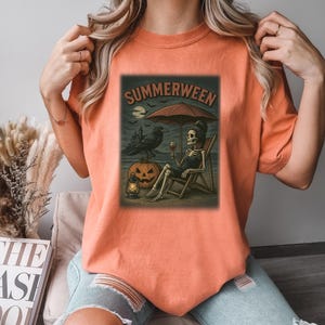 May include: Orange t-shirt with a "Summerween" graphic. The design shows a skeleton on a beach chair under an umbrella, with a raven, jack-o'-lantern, and a lantern. The graphic has a Halloween theme, with a spooky beach scene.