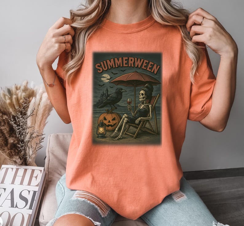 May include: Orange t-shirt with a "Summerween" graphic. The design shows a skeleton on a beach chair under an umbrella, with a raven, jack-o'-lantern, and a lantern. The graphic has a Halloween theme, with a spooky beach scene.