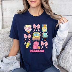May include: Navy blue t-shirt with a colorful school-themed graphic. The design includes books, bows, a school bus, a globe, a backpack, a pencil, and crayons. The name "REBBECA" is printed in pink letters below the graphic.