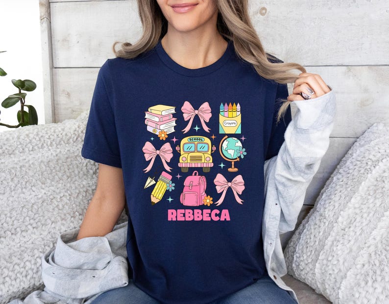May include: Navy blue t-shirt with a colorful school-themed graphic. The design includes books, bows, a school bus, a globe, a backpack, a pencil, and crayons. The name "REBBECA" is printed in pink letters below the graphic.