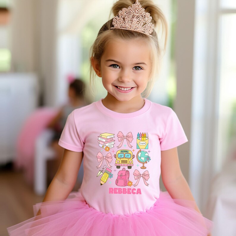 May include: A young girl wearing a pink t-shirt with school-themed graphics, a pink tutu, and a rose gold tiara. The t-shirt features illustrations of books, bows, crayons, a school bus, a globe, a pencil, and a backpack. The name "REBBECA" is printed in pink.