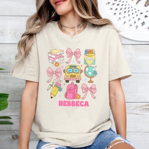 May include: Beige t-shirt with a colorful school-themed graphic. The design includes books, bows, a school bus, a globe, a backpack, a pencil, and a crayon box. The name "REBBECA" is printed in pink. The shirt is a casual, everyday wear item.