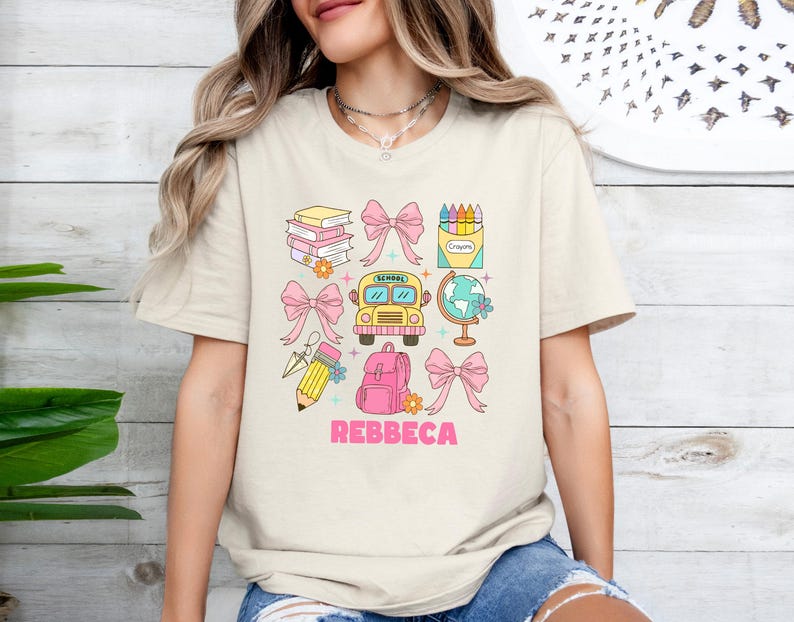 May include: Beige t-shirt with a colorful school-themed graphic. The design includes books, bows, a school bus, a globe, a backpack, a pencil, and a crayon box. The name "REBBECA" is printed in pink. The shirt is a casual, everyday wear item.