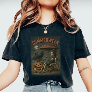 May include: Dark grey t-shirt with a "Summerween" graphic. The illustration shows a skeleton on a beach chair, holding wine, under an umbrella, with a raven, pumpkin, and moon. The shirt has a vintage, worn appearance. The design has a Halloween theme.