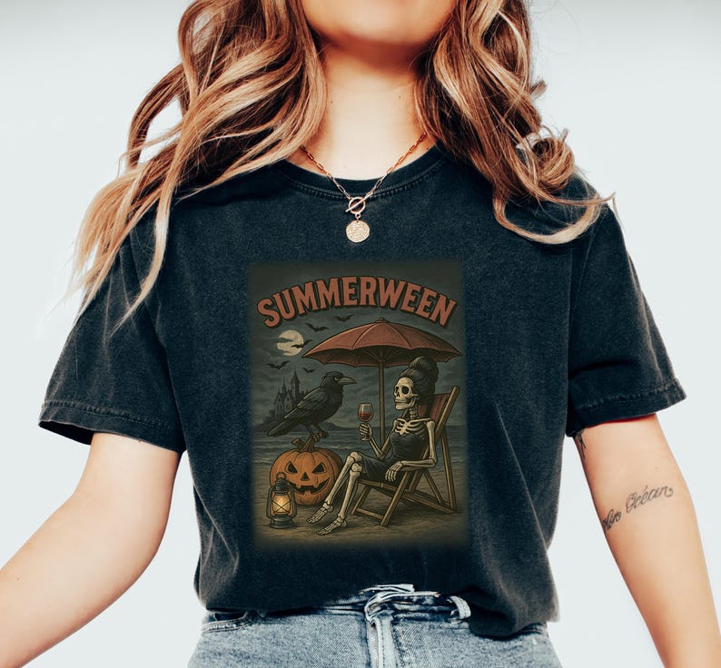 May include: Dark grey t-shirt with a "Summerween" graphic. The illustration shows a skeleton on a beach chair, holding wine, under an umbrella, with a raven, pumpkin, and moon. The shirt has a vintage, worn appearance. The design has a Halloween theme.