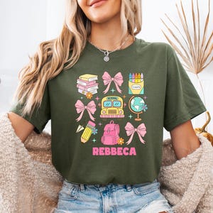 May include: Olive green t-shirt with a colorful school-themed graphic. The design includes books, bows, a school bus, a globe, a backpack, and a pencil. The name "Rebbeca" is printed in pink below the graphic.