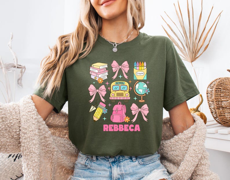 May include: Olive green t-shirt with a colorful school-themed graphic. The design includes books, bows, a school bus, a globe, a backpack, and a pencil. The name "Rebbeca" is printed in pink below the graphic.