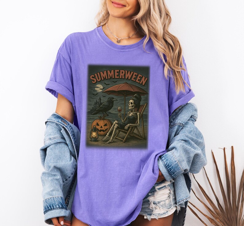 May include: A lilac t-shirt with a graphic that says "Summerween". The graphic shows a skeleton relaxing on a beach chair under a parasol, with a raven, jack-o'-lantern, and a lantern. The shirt is made of a soft material.