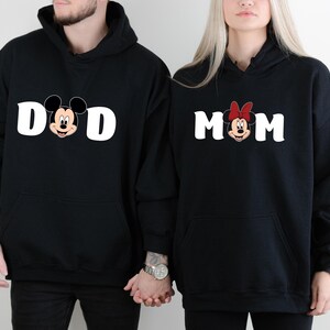 Pu&ograve; includere: Felpe con cappuccio nere con testo bianco e facce di personaggi dei cartoni animati. Una felpa dice "DAD" con Topolino, l'altra dice "MOM" con Minnie. Entrambe hanno una tasca frontale e un cappuccio.