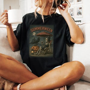 May include: Black t-shirt with a "Summerween" graphic. The graphic shows a skeleton relaxing on a beach chair under an umbrella, with a raven, jack-o'-lantern, and a spooky castle in the background. The skeleton is holding a glass of wine.
