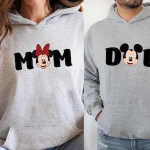 Pu&ograve; includere: Due felpe con cappuccio grigio chiaro. Una dice "MOM" in lettere nere con la faccia di Minnie Mouse e un fiocco rosso. L'altra dice "DAD" in lettere nere con la faccia di Topolino. Entrambe hanno una tasca frontale e un cappuccio.