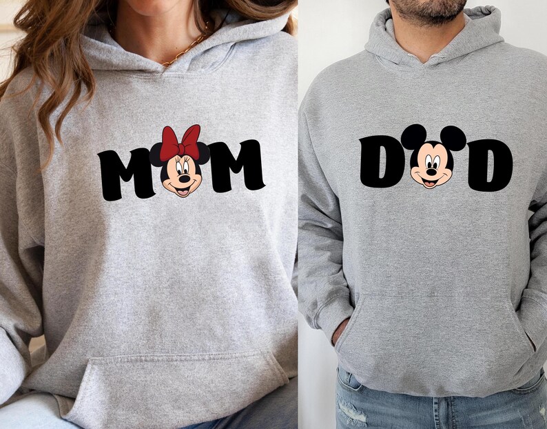 Pu&ograve; includere: Due felpe con cappuccio grigio chiaro. Una dice "MOM" in lettere nere con la faccia di Minnie Mouse e un fiocco rosso. L'altra dice "DAD" in lettere nere con la faccia di Topolino. Entrambe hanno una tasca frontale e un cappuccio.