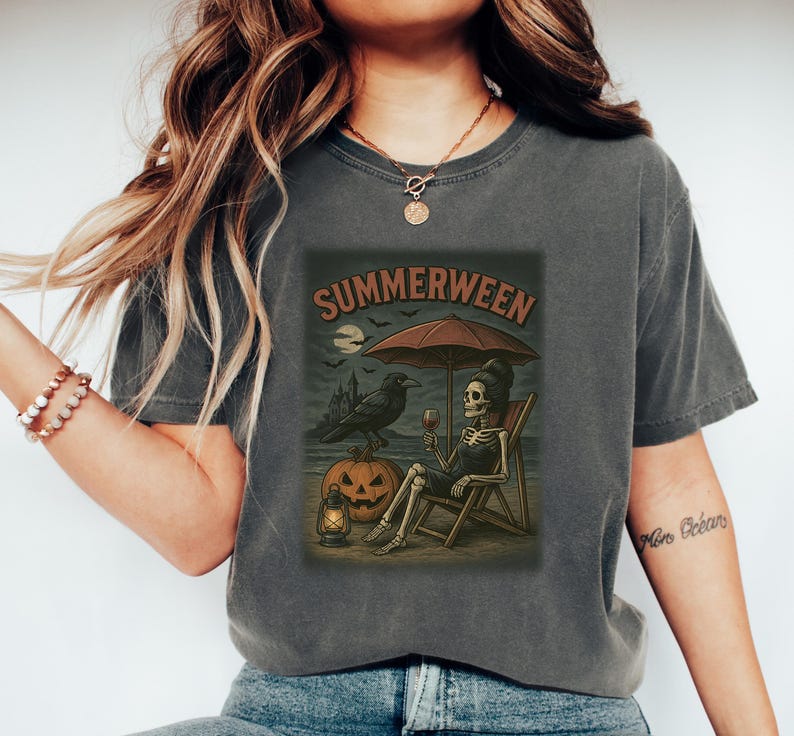 May include: Dark grey t-shirt with a graphic depicting a skeleton on a beach, under an umbrella, with a raven, pumpkin, and the text "SUMMERWEEN". The design has a vintage, Halloween theme.