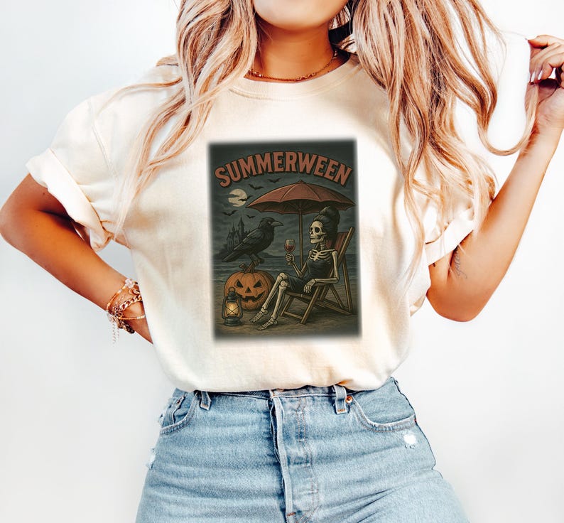 May include: A cream-coloured t-shirt with a graphic that reads "SUMMERWEEN". The design shows a skeleton relaxing on a beach chair under an umbrella, with a raven, pumpkin, and lantern. The graphic has a vintage, spooky aesthetic.