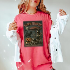 May include: A coral t-shirt with a graphic depicting a skeleton on a beach chair under an umbrella, holding a glass of wine. The image includes a raven, a jack-o'-lantern, and the word "SUMMERWEEN".