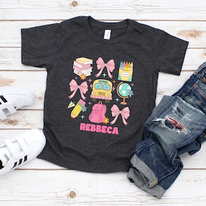 May include: Dark gray t-shirt with a colorful school-themed graphic. The design includes a yellow school bus, pink bows, books, a globe, a backpack, and the name "REBBECA" in pink. The shirt is paired with blue jeans and white sneakers.