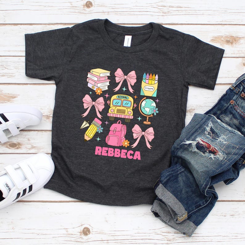 May include: Dark gray t-shirt with a colorful school-themed graphic. The design includes a yellow school bus, pink bows, books, a globe, a backpack, and the name "REBBECA" in pink. The shirt is paired with blue jeans and white sneakers.
