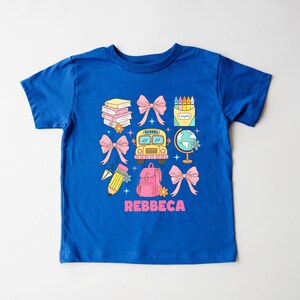 May include: A blue t-shirt featuring a colorful school-themed graphic. The design includes a yellow school bus, pink bows, a globe, books, a pencil, a backpack, and a box of crayons. The name "REBBECA" is printed in pink.