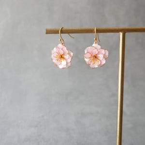 以下が含まれることがあります： 繊細な花のイヤリングのペア。各イヤリングは、金色の縁取りと黄色の中心を持つピンクの花を特徴としています。イヤリングは金色のフックから吊り下げられています。背景は無地のグレーです。