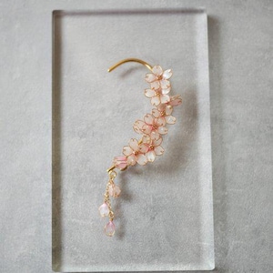 Pode incluir: Um brinco de aro dourado com uma corrente delicada e detalhes de flores cor-de-rosa. As flores são feitas de resina e têm uma aparência translúcida.