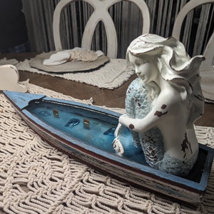 May include: A distressed, white and blue wooden boat with a mermaid figurine. The mermaid has white hair and a blue tail. The boat's interior is blue with small fish and treasure chests.