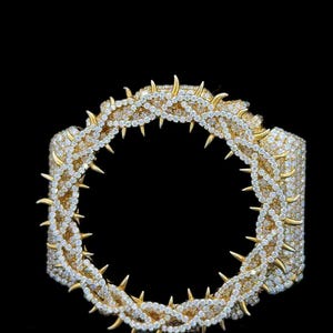 May include: A gold-toned bracelet with a circular design, featuring a band of sparkling white stones and gold-colored spikes. The jewelry is set against a black background, highlighting its intricate details.