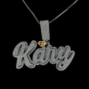Puede incluir: Un collar de cadena de plata con un colgante que dice "Kany" en letra cursiva. El colgante está cubierto de pequeñas piedras transparentes.