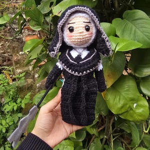 May include: A handmade crochet doll of a nun, dressed in a black habit with a white collar and veil. The doll has a neutral expression and is holding a gray object. The doll is set against a backdrop of green foliage.