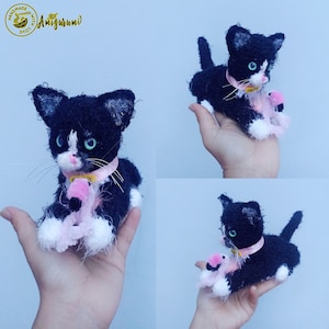 May include: Black and white crocheted cat amigurumi holding a pink flamingo amigurumi. The cat is wearing a pink ribbon around its neck. The amigurumi is handmade with love.