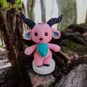 Pode incluir: Um brinquedo de amigurumi de crochê rosa com chifres pretos, asas e detalhes turquesa. O brinquedo está em pé sobre uma base branca.