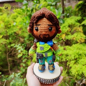 May include: Crocheted amigurumi figure of a person with brown hair, a beard, and a green and blue outfit. The figure is standing on a small round base with a sparkly design.