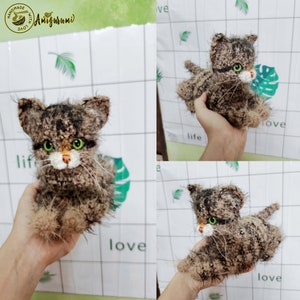 May include: A brown and black crocheted cat with green eyes. The cat is sitting on a hand and is looking at the camera.