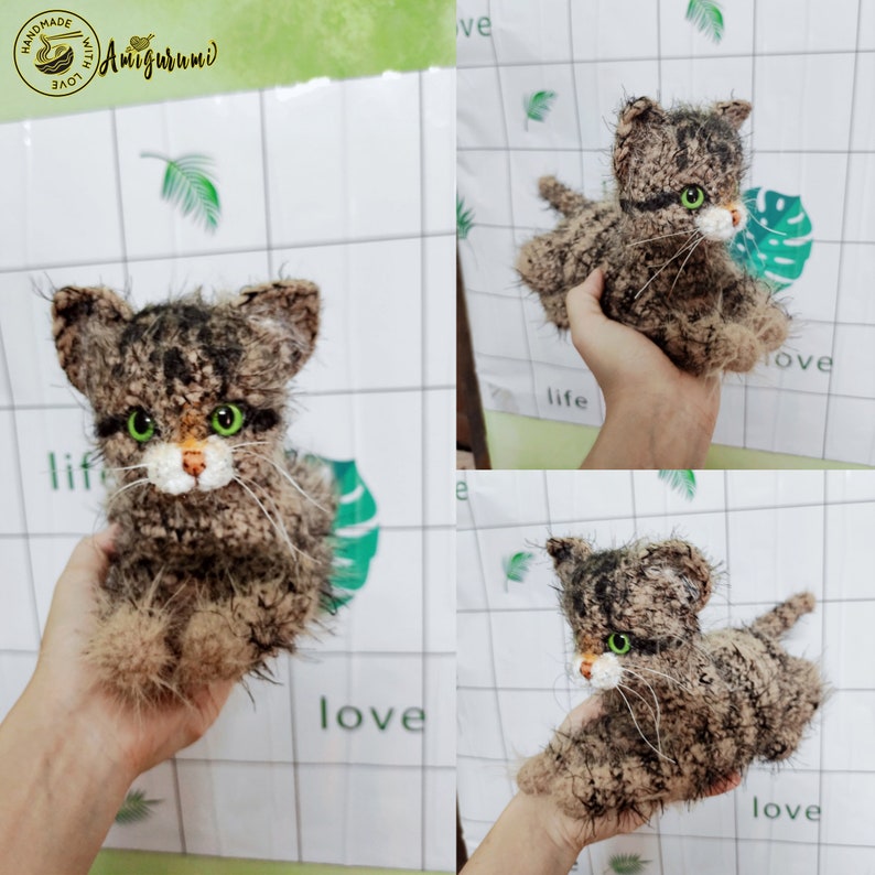 May include: A brown and black crocheted cat with green eyes. The cat is sitting on a hand and is looking at the camera.