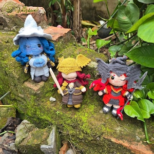 May include: Three crocheted amigurumi dolls. One doll has blue hair and a white hat, another has red hair and a yellow helmet, and the third has red and black hair. The dolls are placed on a moss-covered brick.
