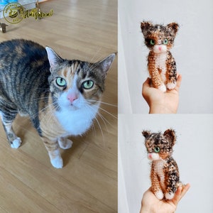 May include: A calico cat with green eyes and a white chest is standing on a wooden floor. Next to the cat is a crocheted calico cat with green eyes and a white chest. The crocheted cat is being held in a hand. The image includes the text "Handmade with Love Amigurumi".
