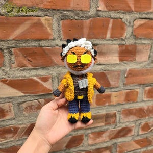 May include: Crocheted amigurumi toy of a person with a white beard, yellow jacket, blue jeans, and yellow sunglasses. The toy is being held in a person's hand.